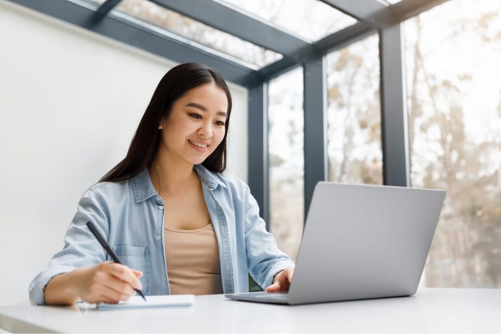 Smiling woman studying online, taking notes while using a laptop in a bright room